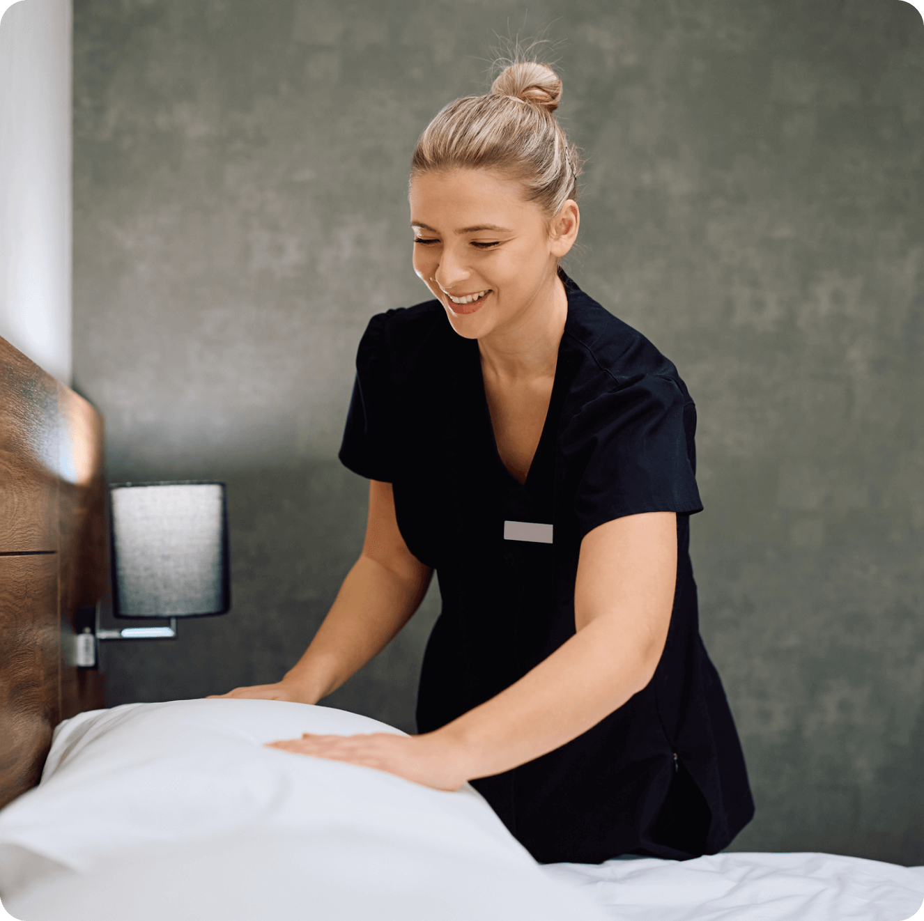 A person in a black uniform is making a bed, smoothing out a white pillow, with a light-colored wall and a lamp in the background.