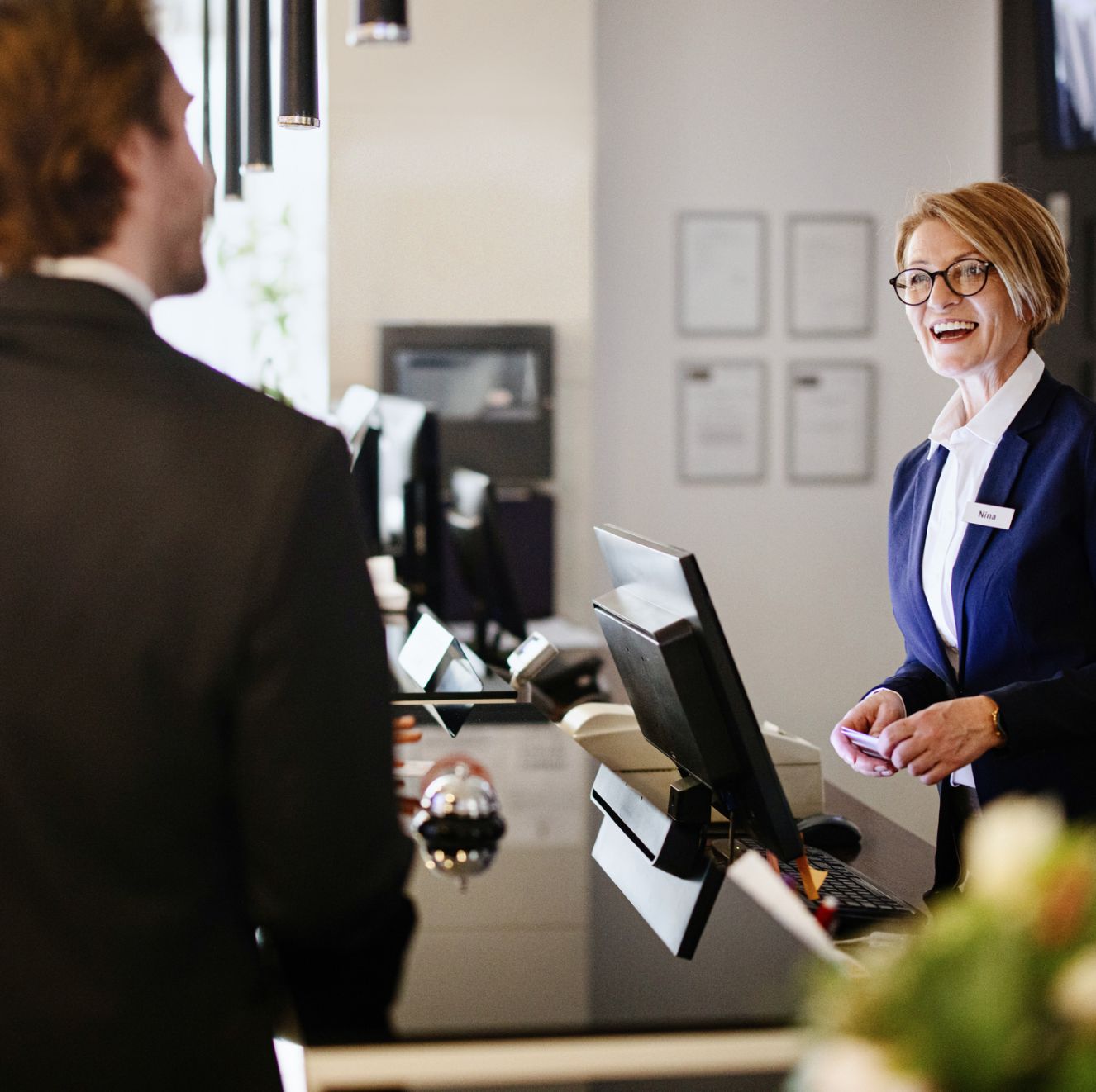 A woman in a blazer smiles at a man across a reception desk in an office setting.