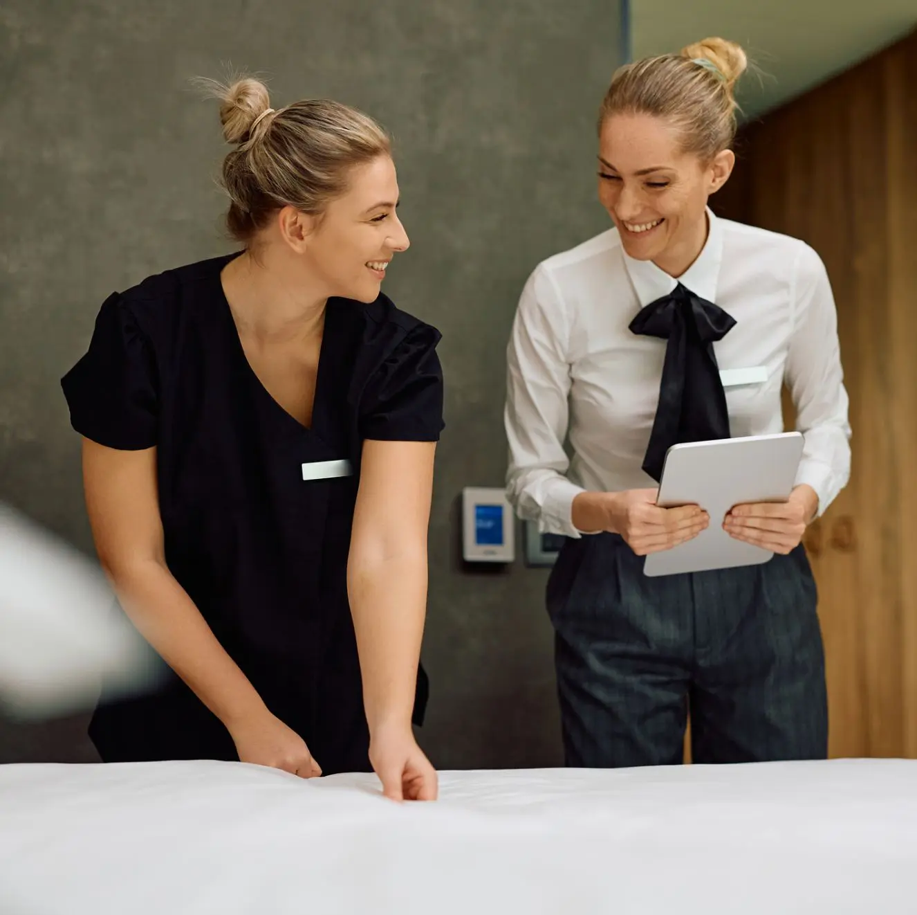 Two women in a hotel room: one is adjusting a bed, and the other is holding a tablet. Both appear to be wearing uniforms.