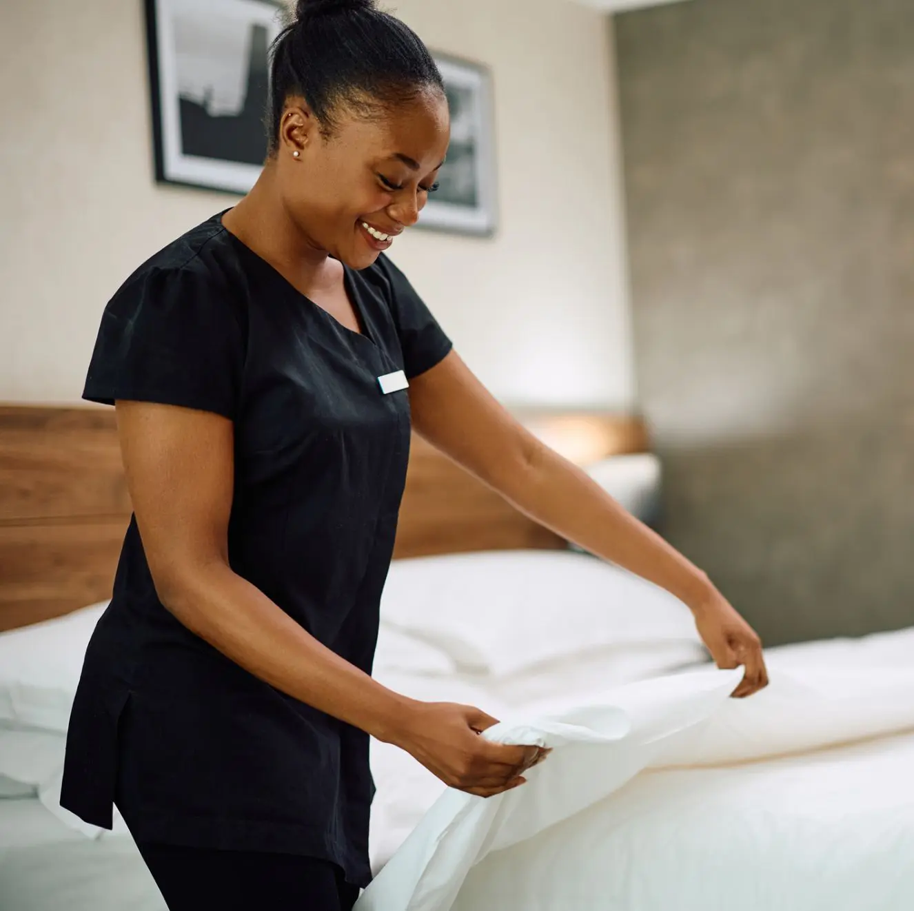 A woman in a black uniform is making a bed, smoothing a white sheet in a well-lit room.