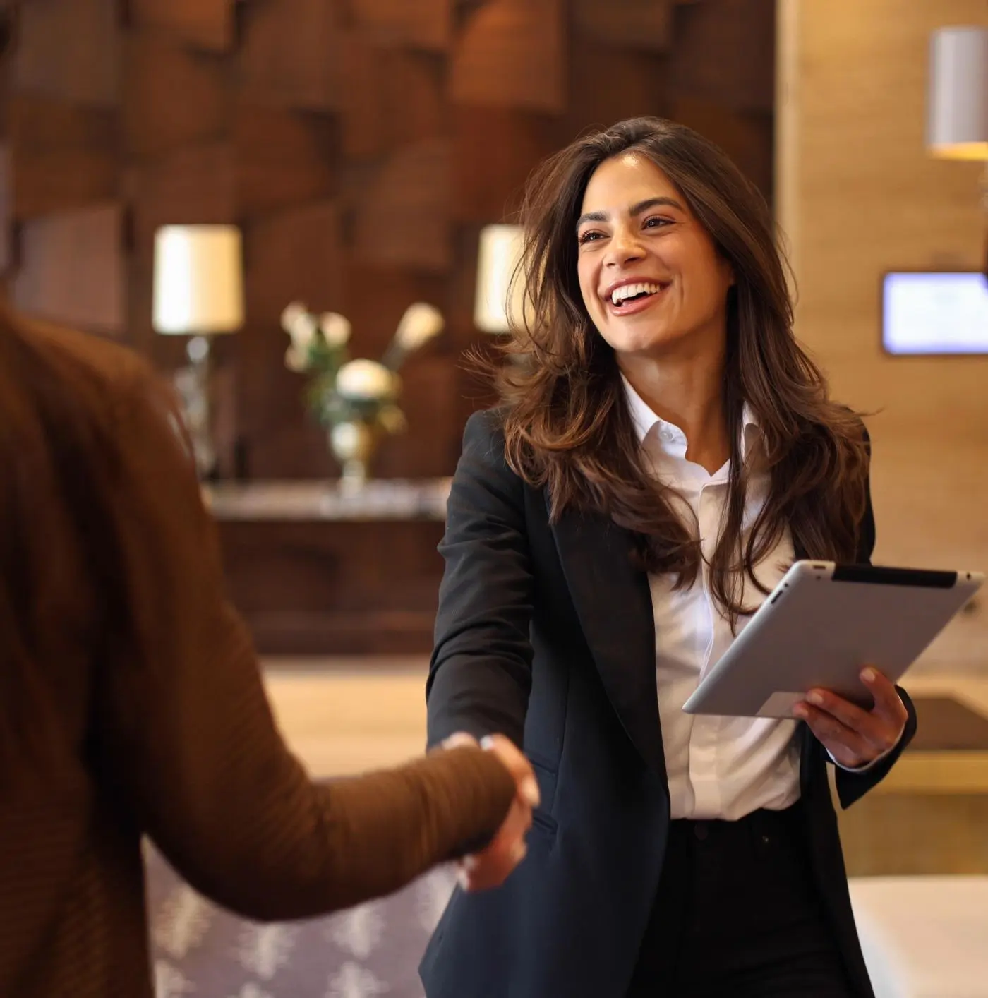 A woman in a business suit smiles and shakes hands while holding a tablet in a modern office setting.