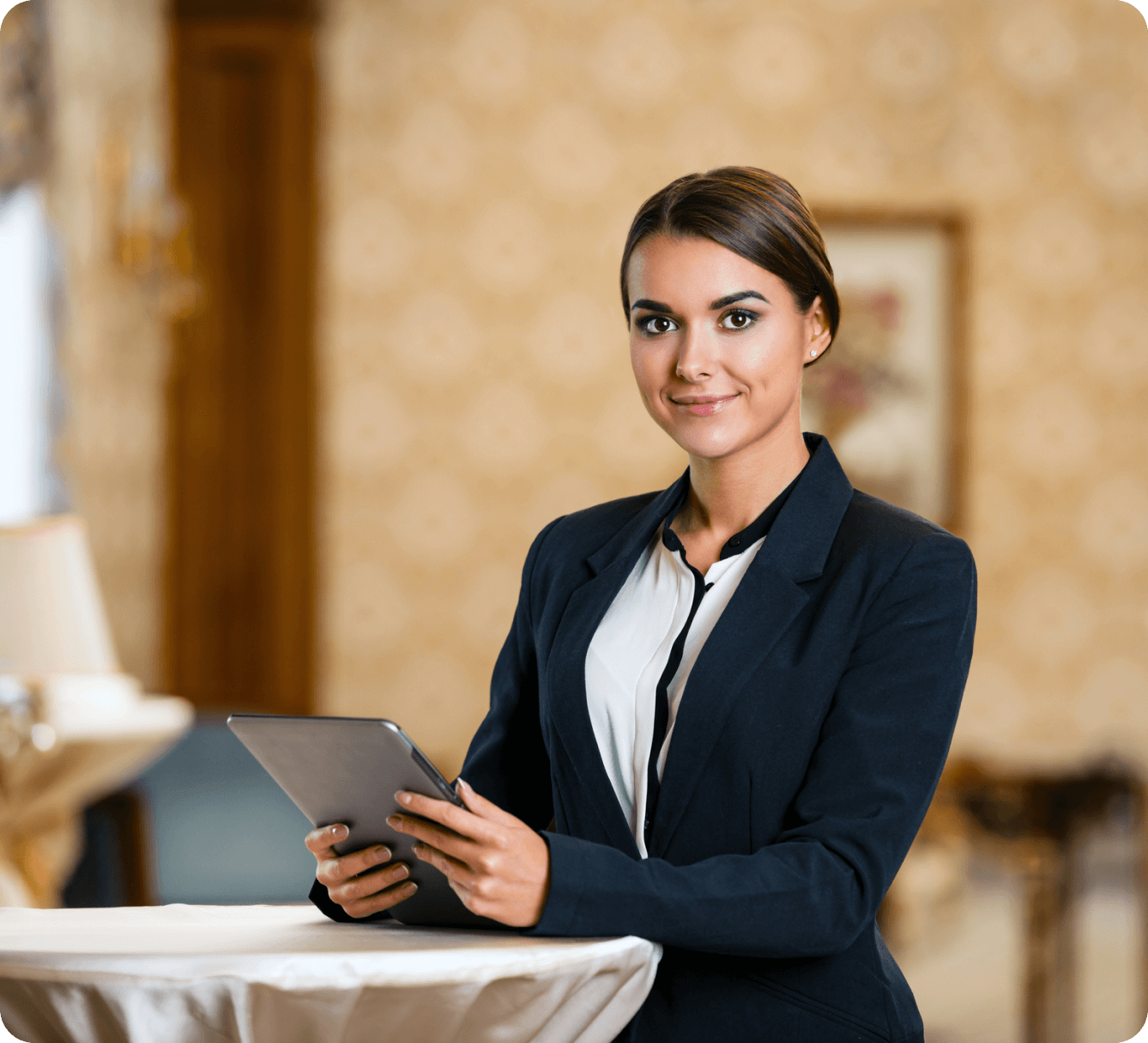 A woman in a business suit stands at a round table holding a tablet, with a blurred interior background.