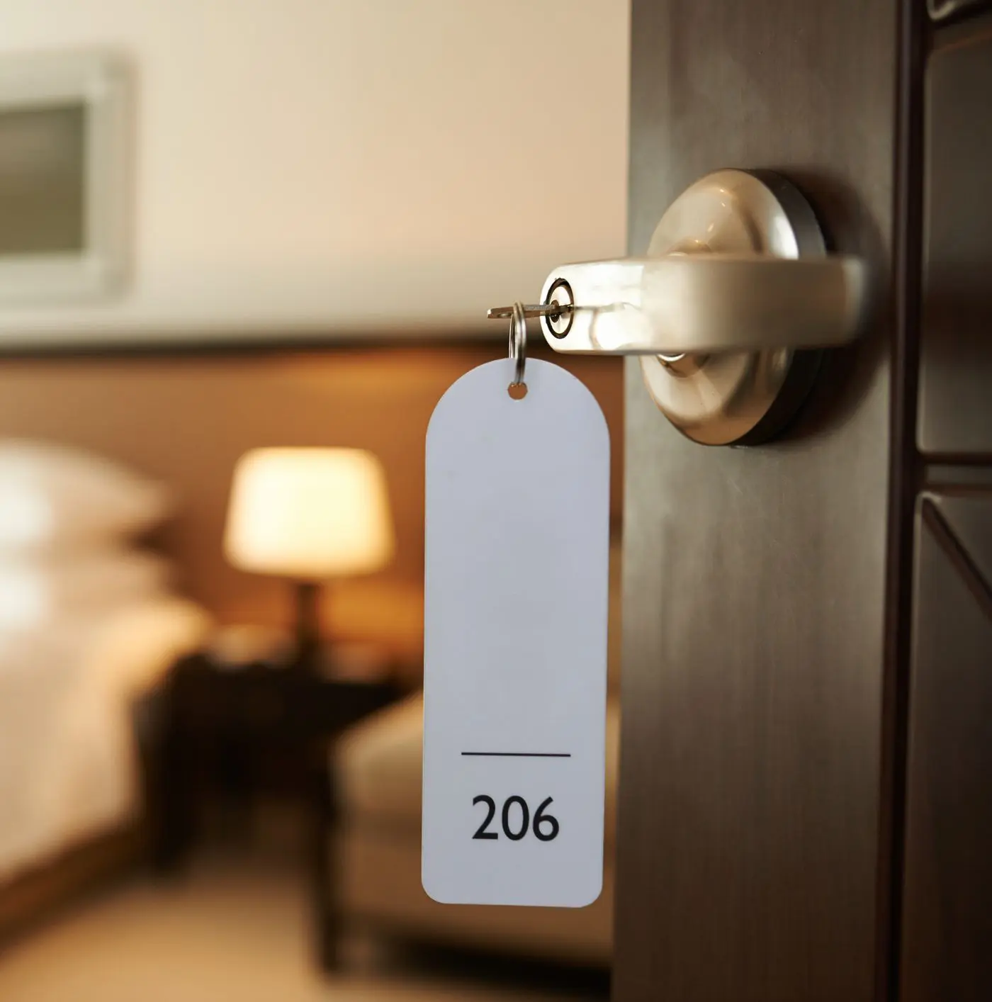 A hotel room door slightly open with a key card and tag labeled "206," revealing a blurred view of a lamp and bed inside.