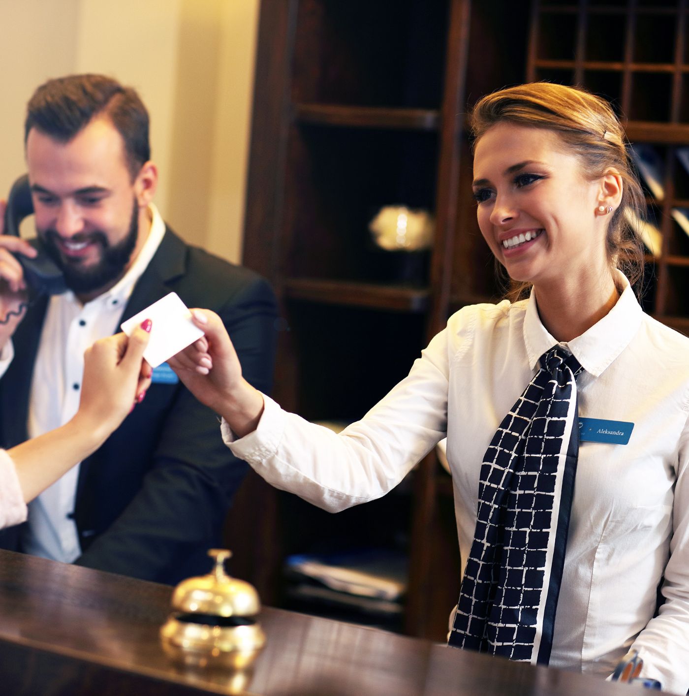 A smiling hotel receptionist hands a key card to a guest, while a male colleague is on the phone behind the desk.