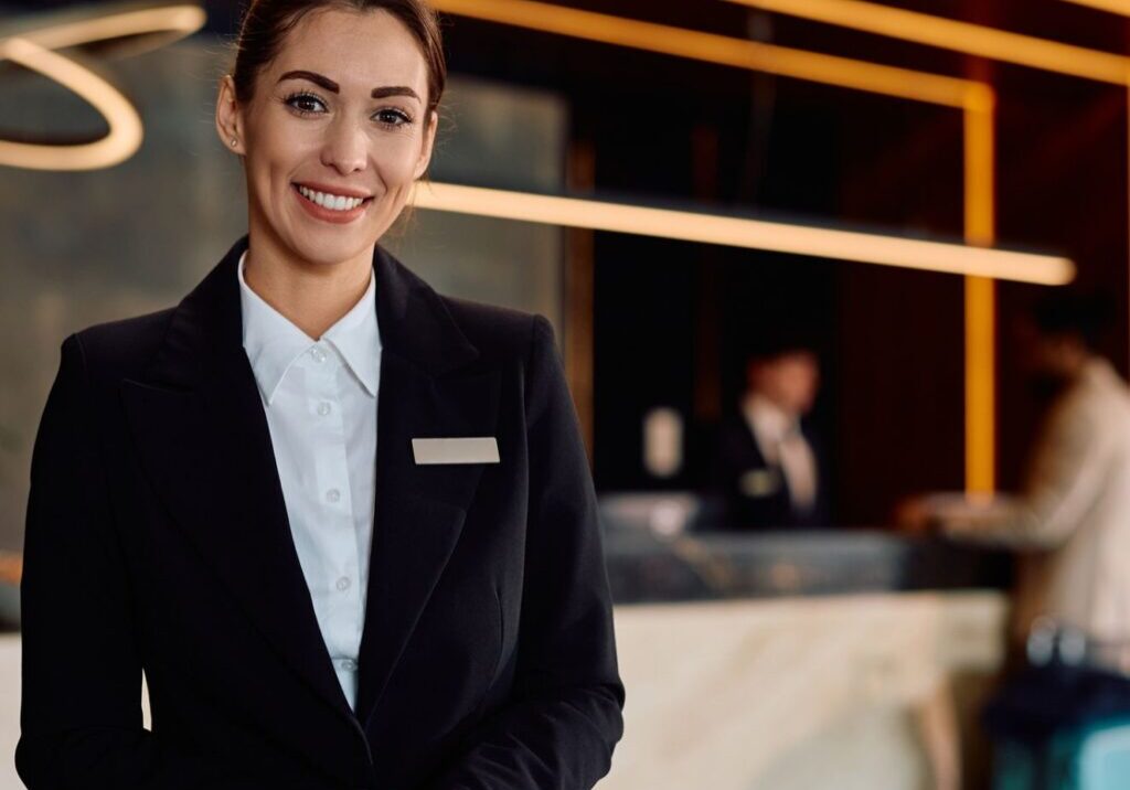 A hotel receptionist in a black suit smiles at the camera, standing in front of a reception desk with two people in the background.